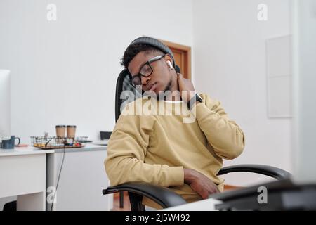 Young African American man in casualwear, eyeglasses and earphones doing relaxing exercise for neck by his workplace in office Stock Photo