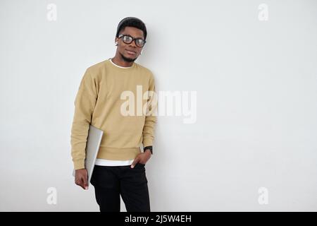 Young contemporary programmer or student in casualwear holding laptop while standing by wall of office and looking at camera Stock Photo