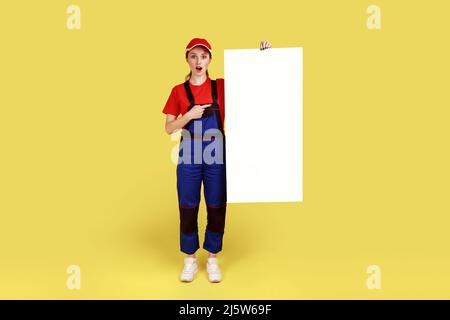 Full length portrait of astonished worker woman holding and pointing at white paper placard with empty space for advertisement or promotional text. Indoor studio shot isolated on yellow background. Stock Photo