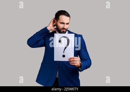 Confused pensive bearded man holding question mark, finding smart solution, asking for advice, looking at camera, wearing official style suit. Indoor studio shot isolated on gray background. Stock Photo