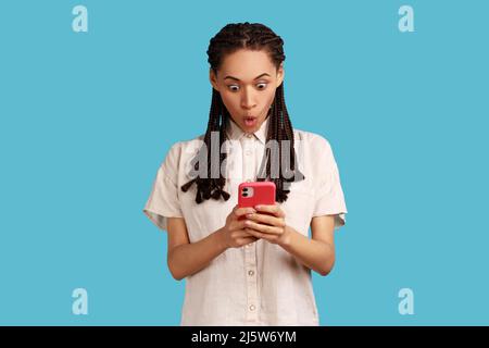 Emotional surprised woman with black dreadlocks stares at smartphone screen, reads amazing news with astonished face, being stunned by something. Indoor studio shot isolated on blue background. Stock Photo