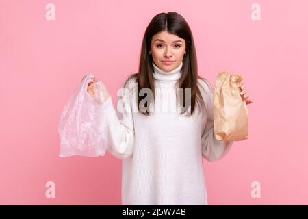 Brunette young woman holding in hands paper and poly bag, needs to make right choice to save planet, wearing white casual style sweater. Indoor studio shot isolated on pink background. Stock Photo