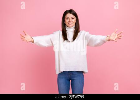 Portrait of happy friendly woman outstretching hands to embrace, giving free hugs and welcoming, wearing white casual style sweater. Indoor studio shot isolated on pink background. Stock Photo