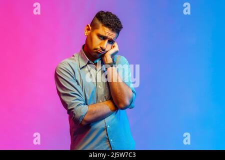 Portrait of depressed man in shirt looking at camera with unhappy sad expression, tired of being lonely, feels bored. Indoor studio shot isolated on colorful neon light background. Stock Photo