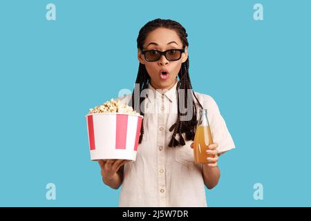 Extremely astonished woman with dreadlocks in 3d imax glasses watching movie film, hold popcorn and drink, watching amazing movie, wearing white shirt. Indoor studio shot isolated on blue background. Stock Photo