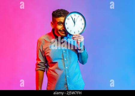 Portrait of funny positive man in shirt standing covering half of face with big wall clock, looking at camera with positive expression. Indoor studio shot isolated on colorful neon light background. Stock Photo