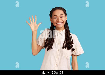 Cheerful friendly woman with black dreadlocks waving palm in hello gesture, meets someone at street, smiles positively, wearing white shirt. Indoor studio shot isolated on blue background. Stock Photo