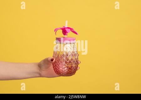 Closeup side view shot of woman hand holding pink glass jar mug with straw for cocktail or smoothie. Indoor studio shot isolated on yellow background. Stock Photo