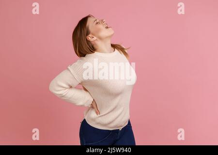 Portrait of blond woman screaming in acute pain and holding sore back, risk of kidney stones disease, pinched nerve, wearing white sweater. Indoor studio shot isolated on pink background. Stock Photo