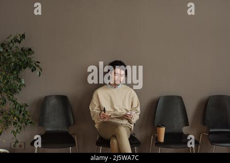 Young man sitting on chair in hallway and reading his resume while waiting for interview Stock Photo