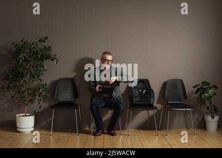 Mature man in eyeglasses checking time on his watch while sitting in waiting room and filling form Stock Photo