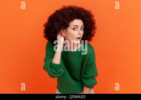 Woman with Afro hairstyle wearing green casual style sweater keeping hand near ear to listen better, having hearing problems, difficult to understand. Indoor studio shot isolated on orange background. Stock Photo