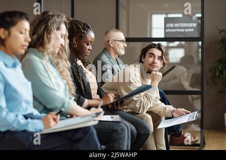 Young man sitting with resume in waiting room among other candidates and waiting for his turn for job interview Stock Photo