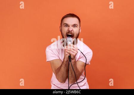 Portrait of angry crazy bearded man holding microphone in hands, looking at camera and screaming loudly, wearing pink T-shirt. Indoor studio shot isolated on orange background. Stock Photo