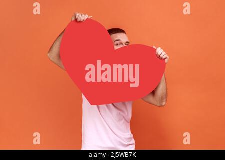 Portrait of man peeping from big red heart and looking at camera with curious prying eyes, feeling affection fondness, wearing pink T-shirt. Indoor studio shot isolated on orange background. Stock Photo