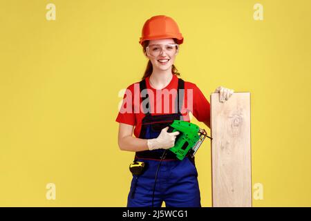 Portrait of satisfied smiling carpenter woman working with fretsaw, holding wooden panel, wearing overalls and protective helmet. Indoor studio shot isolated on yellow background. Stock Photo