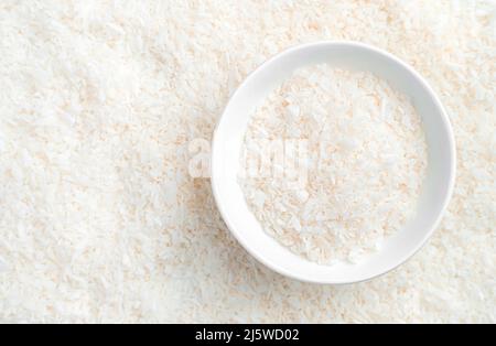 Small coconut chips in the bowl and on the background. Top view, close-up. Stock Photo