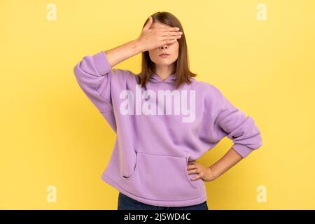 Don't want to look. Portrait of woman keeping hand on eyes and avoiding watch at something shameful, gross, wearing purple hoodie. Indoor studio shot isolated on yellow background. Stock Photo