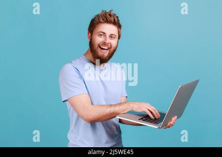 Portrait of smiling bearded man holding laptop in hand and typing, blogger making posts in social networks, chatting with followers, looking at camera. Indoor studio shot isolated on blue background. Stock Photo