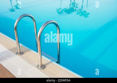 Palm trees are reflected in a pool of turquoise water. Metal ladder for the pool. Stock Photo
