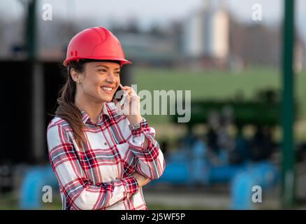 Smiling young female engineer with helmet standing in front of irrigation system plant in agricultural field and talking on her phone. Stock Photo