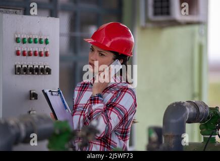 Busy female engineer with helmet and papers talking on the phone in front of electric boar in outdoor agricultural plant Stock Photo