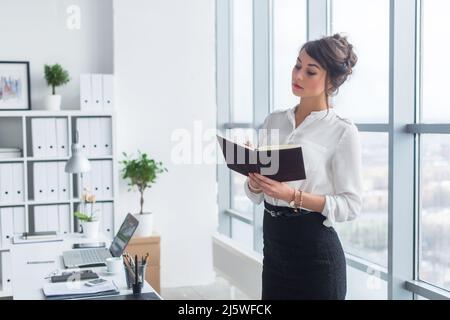 Female office worker writing down notes and day plan in her notebook, standing at workplace, front view portrait Stock Photo