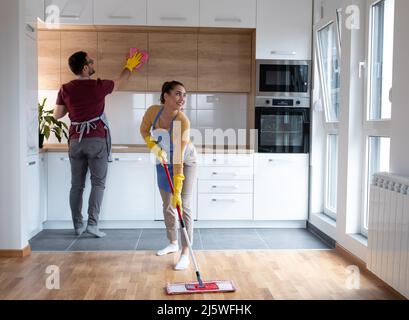 Couple doing chores together while young woman cleaning floor with mop man wipes the dust from kitchen in their apartment. Stock Photo