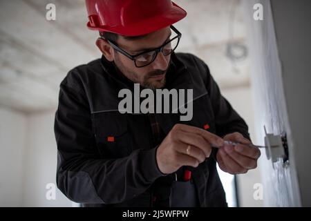 Young male electrician with helmet holding a screwdriver and installing power socket in apartment. Stock Photo