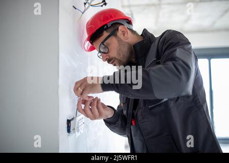 Serious male electrician with helmet installing power socket in the apartment and using screwdriver. Stock Photo