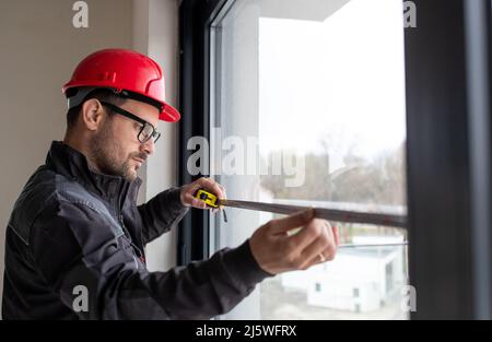 Male worker wearing helmet measuring window with measuring tape for installing blinds indoors. Stock Photo
