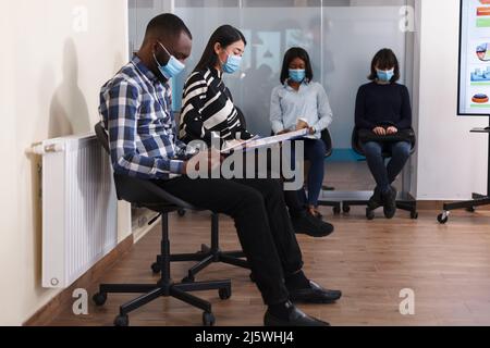 Multi ethnic job applicants sitting in business agency lobby area while waiting for employment manager to show up for interview series. Unemployed people reviewing CV resumes while in waiting room. Stock Photo