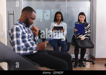Multi racial job applicants sitting in lobby area of business company while reviewing CV resumes before interview series. Tensioned diverse people in waiting for HR department head manager to show up. Stock Photo