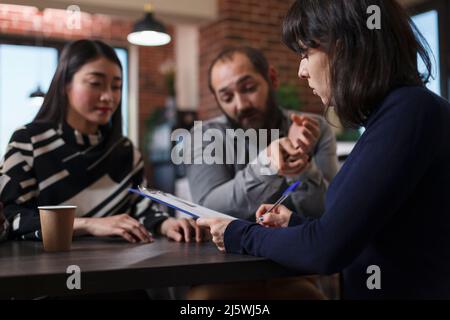 Young adult recruiter sitting at desk in office space while reviewing job applicants CVs. Multiethnic candidates talking with startup company interviewer while talking about career oportunities. Stock Photo