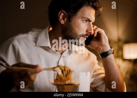 White man eating asian noodles while talking on cellphone at office Stock Photo