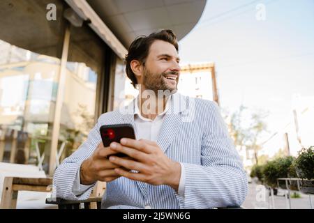 Young man smiling and using mobile phone while sitting at cafe outdoors Stock Photo