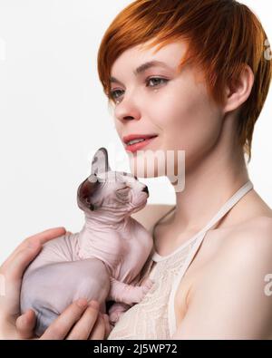 Cute redhead young woman gently hugging sleeping kitten to her chest and looking away. Portrait of pretty woman with short hair. Studio shot on white Stock Photo