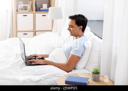 happy indian man with laptop in bed at home Stock Photo