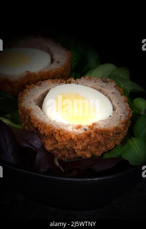 Scotch egg halved on lettuce leaves in a black dish.  On a stone background Stock Photo