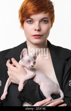 Serious young woman in black jacket holding in hands Canadian Sphynx Cat. Pretty redhead woman 25 years old with short hair looking at camera Stock Photo