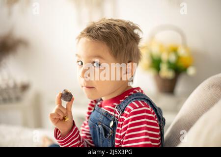 Cute child at home with little newborn chicks, eating chocolate candy in bed, happy kid Stock Photo