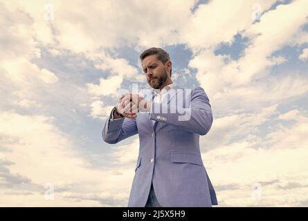 handsome mature man businessman in suit check time on wristwatch on sky background, punctuality. Stock Photo