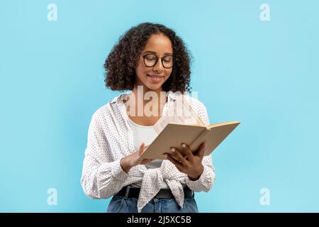 Happy black female student reading textbook, making home assignment, studying for exam on blue studio background Stock Photo