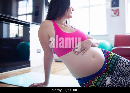 Pregnant woman in sports bra resting on yoga mat at home Stock Photo