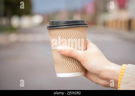 Hand of young woman holding brown paper coffee cup against blurred city background. Mockup image of paper cup for hot beverages, plastic lid. Selectiv Stock Photo