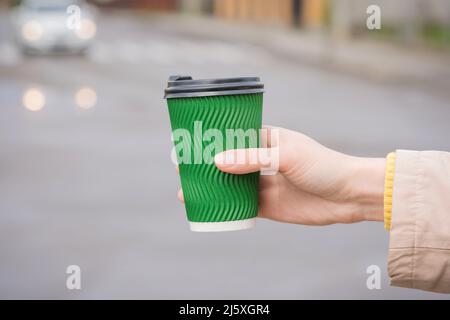 Young woman holding green paper coffee cup against blurred city background. Mockup image of paper cup with plastic lid for hot beverages. Selective fo Stock Photo