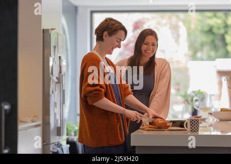 Happy female friends cutting loaf cake in kitchen Stock Photo