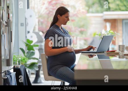 Pregnant woman working from home at laptop in kitchen Stock Photo