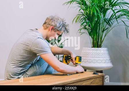 A young man with an electrical screwdriver assembles a tv stand console according to instructions in his new house. Man assembling furniture at home u Stock Photo