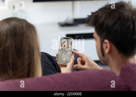 Two parents sitting on the sofa and monitoring their baby on the phone Stock Photo